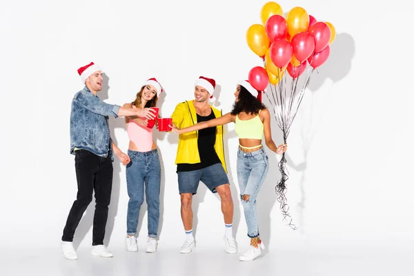 Hommes heureux et filles multiculturelles chapeaux Santa avec des tasses en plastique près de ballons sur blanc — Photo de stock