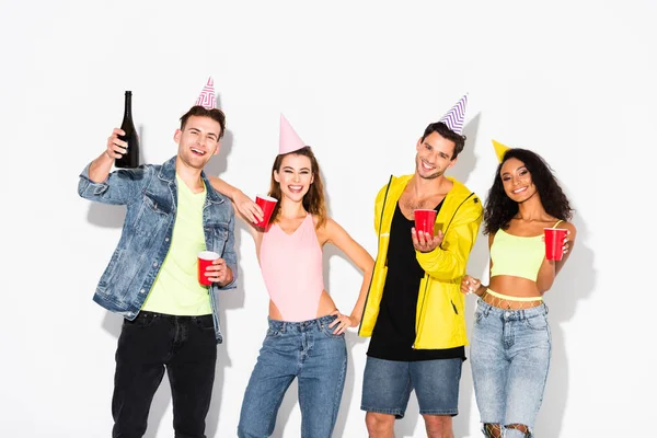 Happy multicultural friends in party caps holding plastic cups on white — Stock Photo