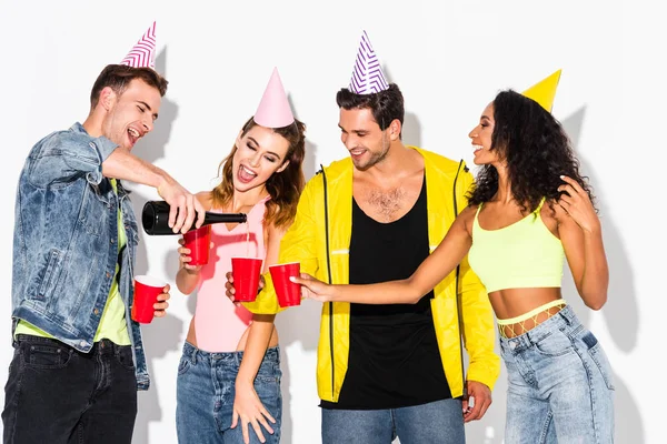 Hombre feliz vertiendo champán en copas de plástico cerca de amigos multiculturales en gorras de fiesta en blanco - foto de stock