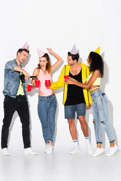 Hombre alegre verter champán en copas de plástico cerca de amigos multiculturales en gorras de fiesta en blanco - foto de stock