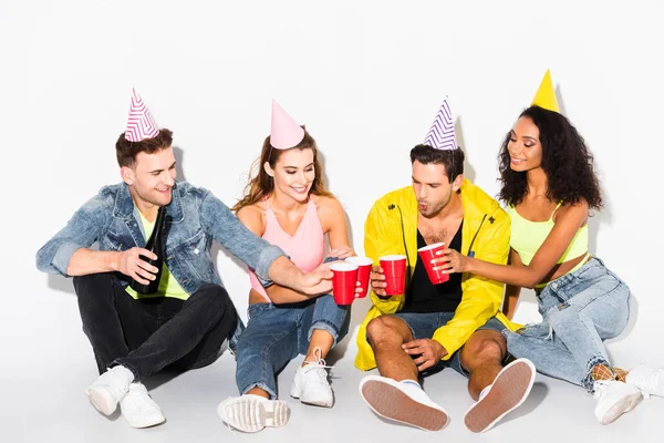 Hombre feliz sosteniendo copas de plástico y tintineo con amigos multiculturales en gorras de fiesta en blanco - foto de stock