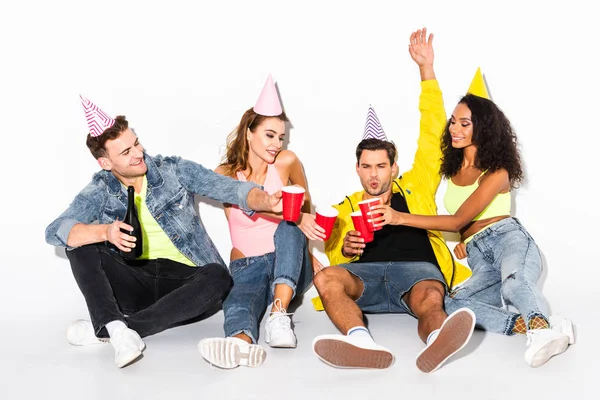 Cheerful man holding plastic cups and clinking with multicultural friends in party caps on white — Stock Photo