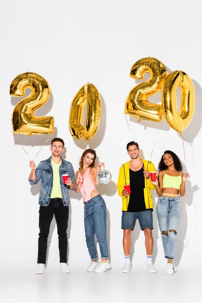 Hombres y mujeres multiculturales felices sosteniendo copas de plástico cerca de globos en blanco - foto de stock