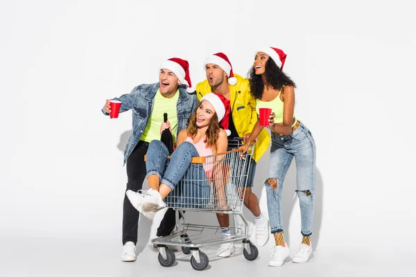 Chica feliz sentado en el carrito de la compra con botella cerca de amigos multiculturales con copas de plástico en blanco - foto de stock