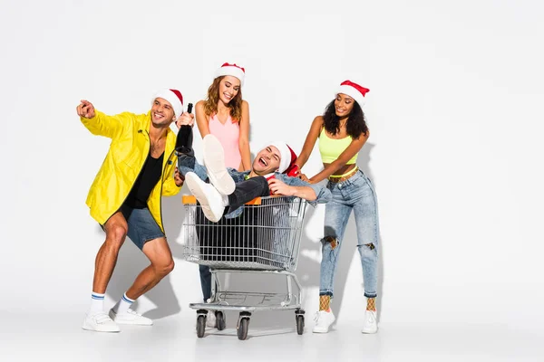 Cheerful man pointing with finger near friend sitting in shopping cart and multicultural girls on white — Stock Photo