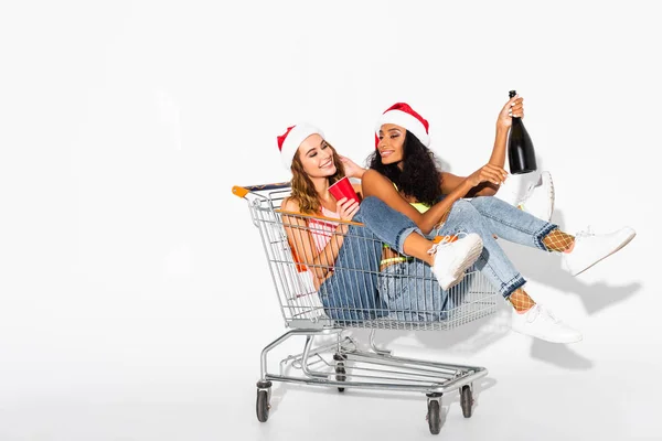 Meninas multiculturais felizes sentadas no carrinho de compras com garrafa de champanhe em branco — Fotografia de Stock