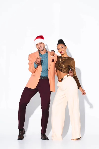 Fröhliche afrikanisch-amerikanische Frau steht mit Mann in Weihnachtsmütze und zeigt mit dem Finger auf weiß — Stockfoto