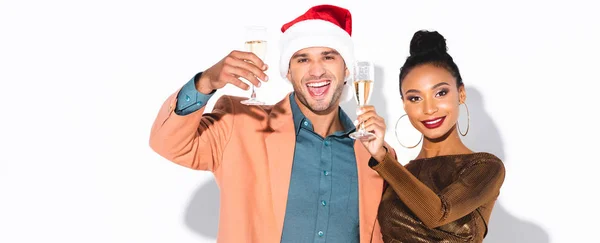 Panoramaaufnahme eines fröhlichen afrikanisch-amerikanischen Mädchens mit Champagnerglas in der Nähe eines Mannes mit Weihnachtsmütze auf weißem Hintergrund — Stockfoto