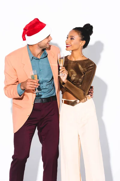 Fröhliches afrikanisch-amerikanisches Mädchen hält Champagnerglas in der Hand und schaut den glücklichen Mann mit Weihnachtsmütze auf weißem Hintergrund an — Stockfoto