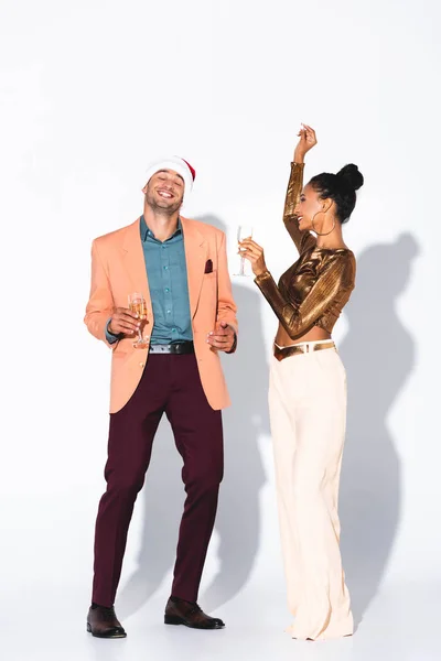 Schöne afrikanisch-amerikanische Mädchen mit Champagnerglas und Blick auf glücklichen Mann mit Weihnachtsmütze auf weiß — Stockfoto