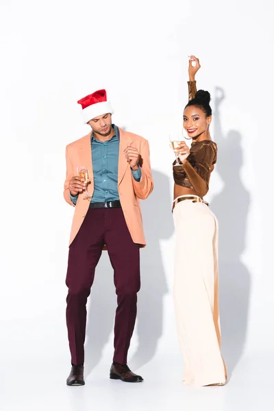 Beautiful african american girl holding champagne glass and looking at camera near man in santa hat on white — Stock Photo