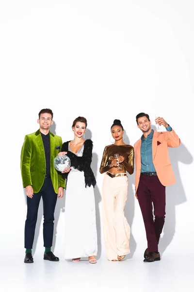 Amigos multiculturales felices y de moda sosteniendo copas de champán y bola disco en blanco - foto de stock
