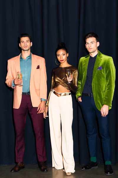 Hermosa mujer afroamericana y hombres guapos sosteniendo copas de champán en azul oscuro - foto de stock