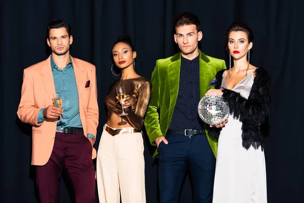 Hermosas mujeres multiculturales y hombres guapos sosteniendo copas de champán en azul oscuro - foto de stock