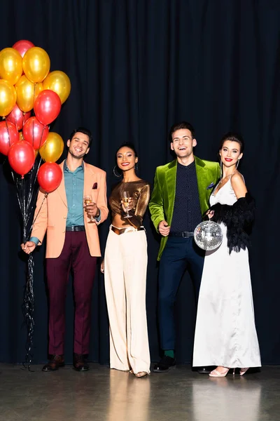 Alegres mujeres multiculturales y hombres guapos sosteniendo copas de champán cerca de globos en azul oscuro - foto de stock