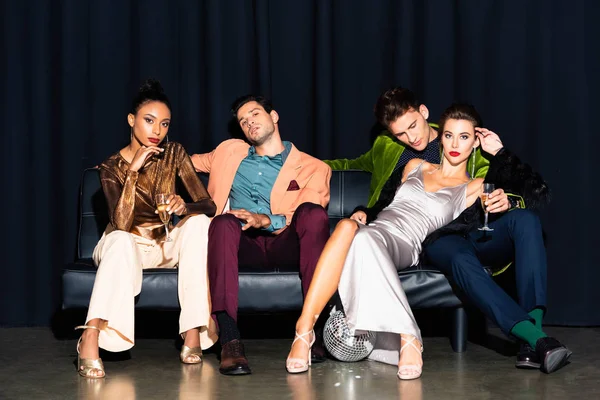 Hermosas mujeres multiculturales y hombres guapos sosteniendo copas de champán mientras se sienta en el sofá en azul oscuro - foto de stock