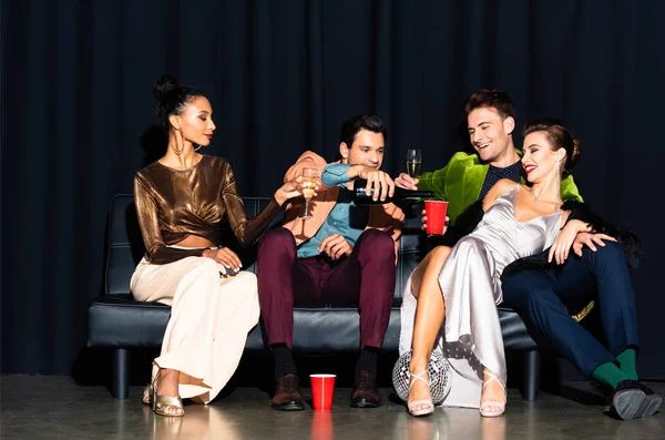 Schöner Mann gießt Champagner in Plastikbecher neben multikultureller Frau, während er auf Sofa auf dunkelblau sitzt — Stockfoto