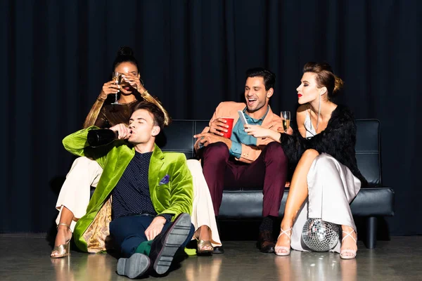 Handsome mna drinking champagne from bottle near multicultural friends sitting on dark blue — Stock Photo