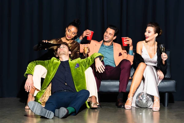 Happy african american girl holding bottle near man sitting on floor near friends on dark blue — Stock Photo