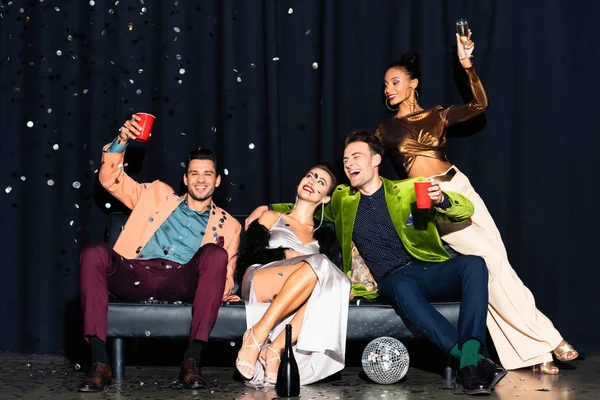 Cheerful multicultural friends sitting on sofa near falling confetti on dark blue — Stock Photo
