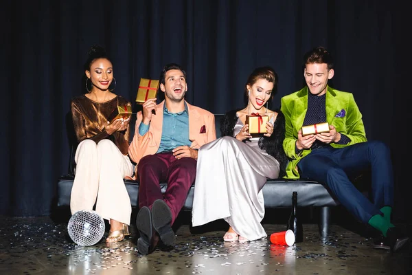 Happy multicultural friends sitting on sofa and holding presents on dark blue — Stock Photo