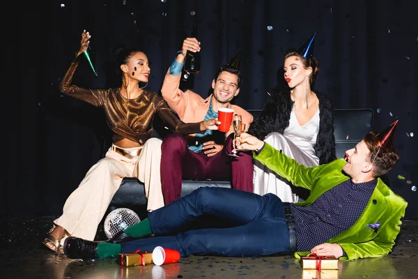 Fröhliche multikulturelle Freunde in Partymützen klirren in der Nähe der Discokugel auf dunkelblau — Stockfoto