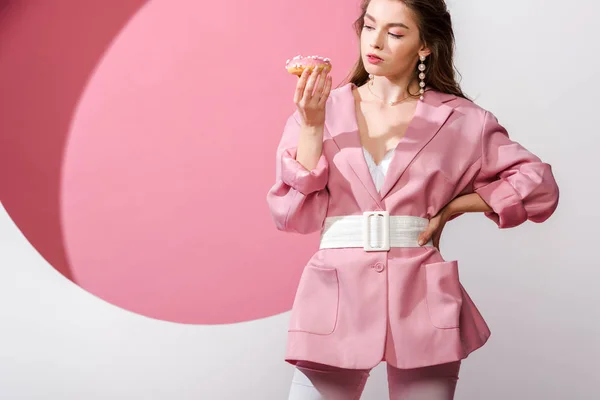 Atractiva mujer de pie con la mano en la cadera y mirando donut dulce en blanco y rosa - foto de stock