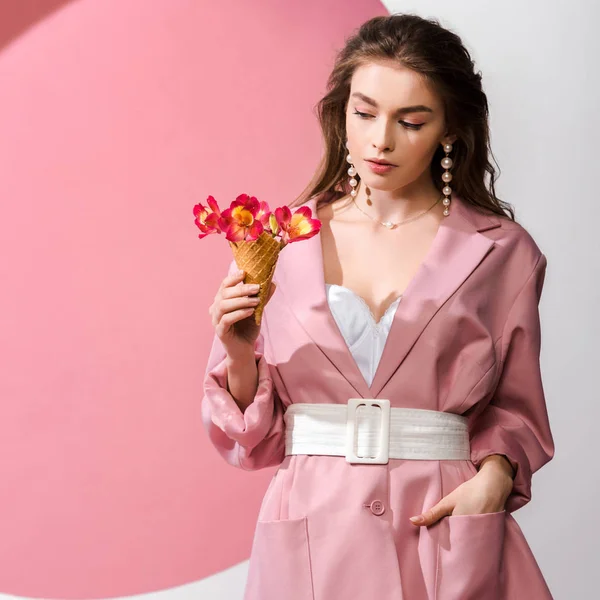 Attraktives Mädchen, das mit der Hand in der Tasche steht und Eiskegel mit Blumen auf weiß und rosa anschaut — Stockfoto