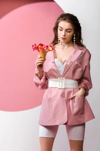 Atractiva mujer de pie con la mano en el bolsillo y mirando el cono de helado con flores en blanco y rosa - foto de stock