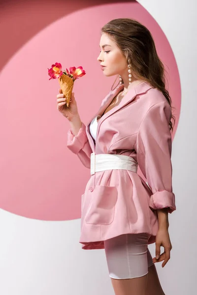 Vue latérale de la femme attrayante regardant le cône de crème glacée avec des fleurs sur blanc et rose — Photo de stock