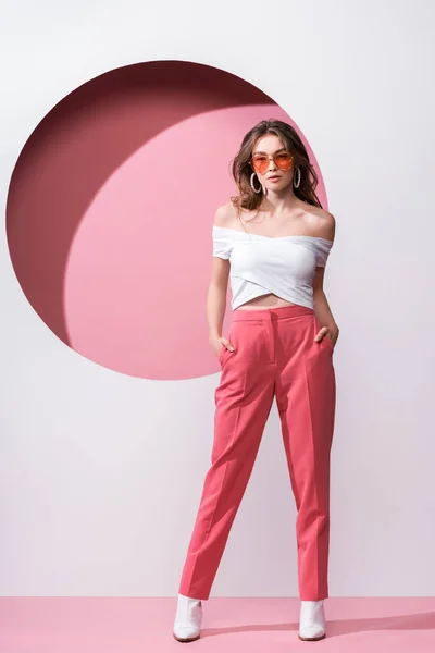Привлекательная девушка в солнечных очках, стоящая с руками в карманах на розовом и белом — стоковое фото