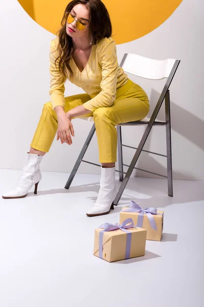 Menina atraente em óculos de sol sentado na cadeira perto de presentes em laranja e branco — Fotografia de Stock