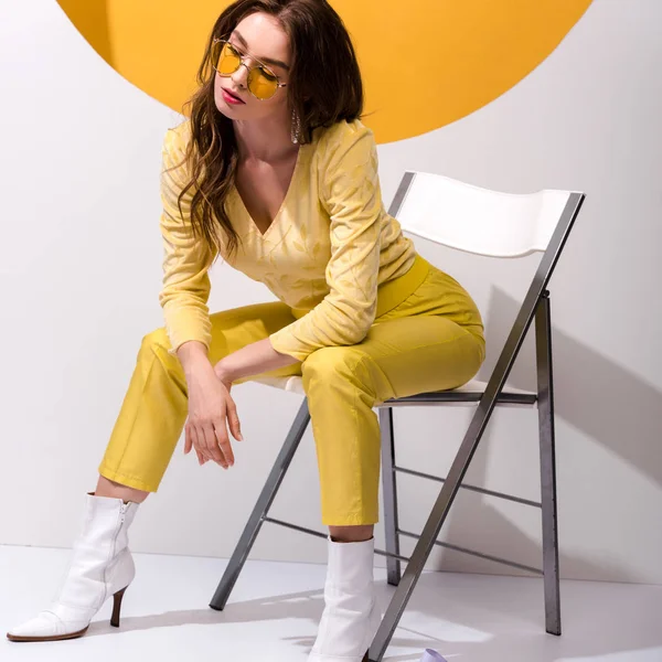 Приваблива дівчина в сонцезахисних окулярах, сидячи на стільці на помаранчевому і білому — стокове фото