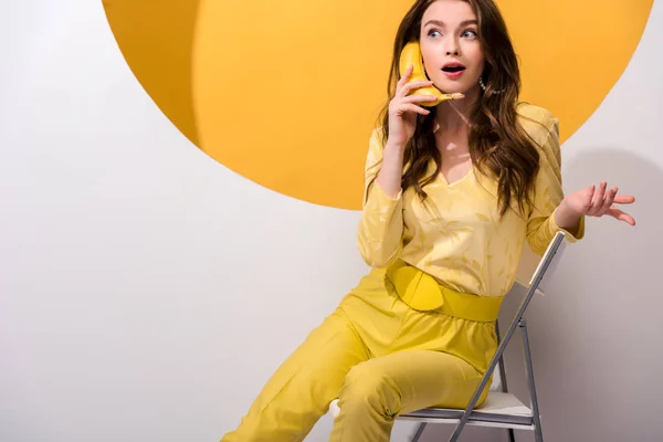 Menina bonita segurando banana enquanto sentado e gesticulando em branco e laranja — Fotografia de Stock