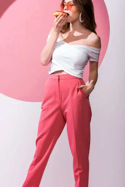 Attraente ragazza mangiare gustosa ciambella e in piedi con mano in tasca su rosa e bianco — Foto stock
