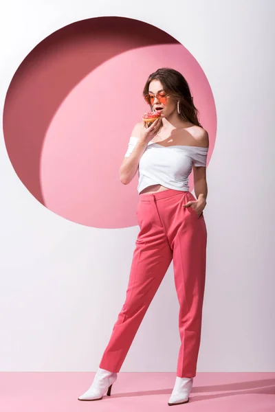 Schönes Mädchen hält leckeren Donut und steht mit der Hand in der Tasche auf rosa und weiß — Stockfoto