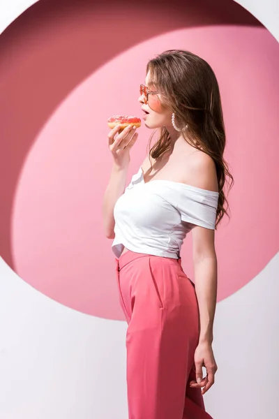 Side view of beautiful girl holding tasty donghnut and standing on pink and white — стоковое фото