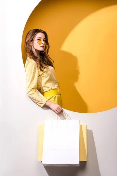 Joven atractiva mujer en gafas de sol sosteniendo bolsas de compras en naranja y blanco - foto de stock