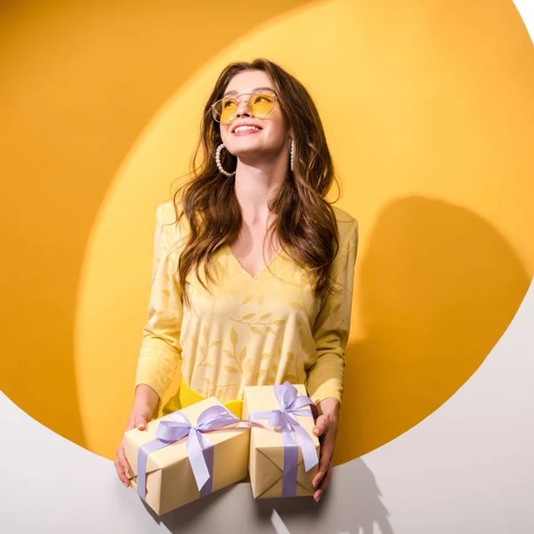 Happy woman in sunglasses holding gifts on orange and white — Stock Photo