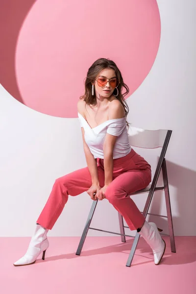 Attrayant femme dans des lunettes de soleil assis sur la chaise sur rose et blanc — Photo de stock