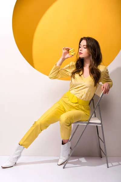 Attractive woman sitting on chair and holding macarons on orange and white — Stock Photo