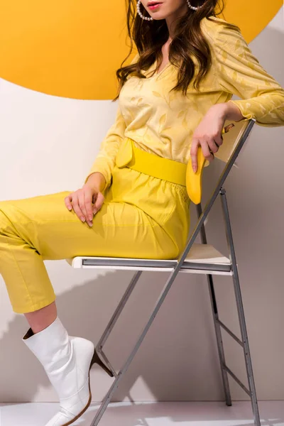Vista ritagliata di giovane donna seduta sulla sedia e che tiene banana su bianco e arancione — Foto stock