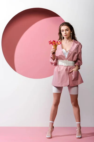 Привлекательная женщина стоит с рукой в кармане и держит мороженое конус с цветами на розовом и белом — стоковое фото