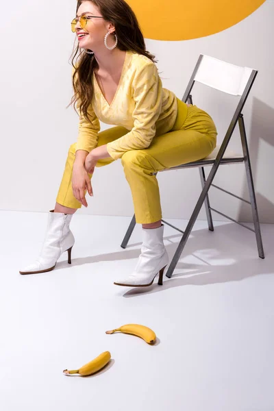 Glückliche Frau lächelt, während sie auf einem Stuhl neben gelben Bananen auf weiß sitzt — Stockfoto