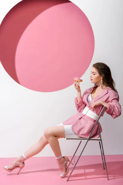 Attractive woman sitting on chair and holding sweet doughnut on pink and white — Stock Photo