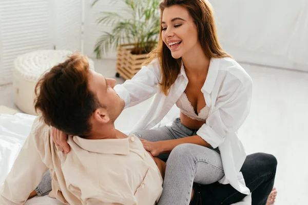 Novia sonriente y atractiva sentado y abrazando novio en el apartamento - foto de stock