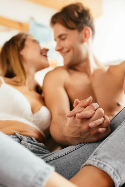Selective focus of smiling boyfriend and girlfriend in bra holding hands in apartment — Stock Photo