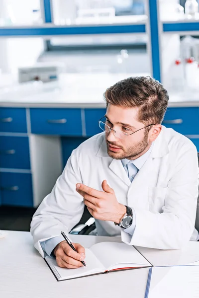 Schöner Arzt mit Brille, der in Notizbuch schreibt und in der Klinik gestikuliert — Stockfoto