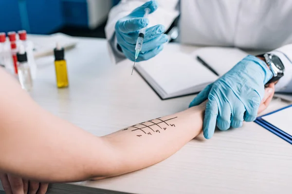 Cropped view of allergist in latex gloves holding syringe near woman while doing allergy test in clinic — Stock Photo