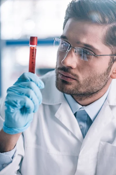 Handsome bearded immunologist in glasses looking at test tube with red liquid — Stock Photo